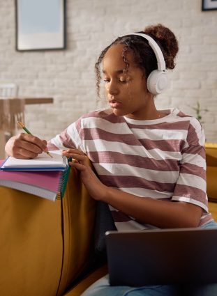 African American teenage girl taking notes while studying over laptop at home.